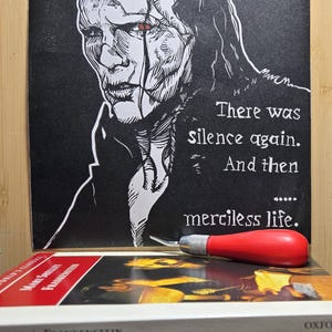 May include: Black and white print of a stylized figure with the text "There was silence again. And then merciless life." A red-handled craft knife rests on a stack of "Frankenstein" books.