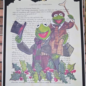 Könnte beinhalten: Ein Kunstwerk mit zwei Kermit-der-Frosch-Charakteren in Kostümen aus "A Christmas Carol", vor dem Hintergrund von Buchseiten. Das Bild enthält Stechpalmen und Beeren am unteren Rand und ist auf schwarzem Papier montiert, mit einer Holzklammer.