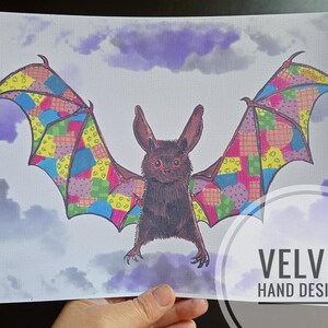 May include: A detailed illustration of a bat with patchwork wings in various colours, set against a cloudy, purple-toned sky. The bat has large ears and a detailed face. The text "VELVET HAND DESIGNS" is visible in the lower right corner.