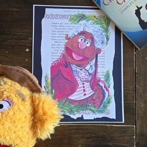 May include: A print featuring a colourful illustration of a puppet character in a red coat over a page from A Christmas Carol by Charles Dickens, accompanied by a copy of the book and a fuzzy yellow puppet with a brown hat in the foreground.