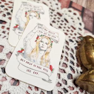 May include: A white card with a hand-drawn illustration of a woman with long blonde hair wearing a crown of red flowers. The woman is holding a banner that reads "The truth will set you free."  A second banner below reads "But first it will piss you off."