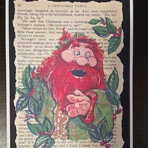 May include: A rectangular print featuring a page from A Christmas Carol with text and a central illustration of a red-bearded figure with a wreath of holly and red berries. The figure has a wide mouth and blue eyes, framed by green leaves.