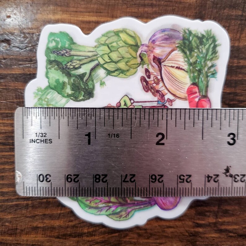 May include: A sticker featuring a watercolor illustration of various vegetables, including an artichoke, onion, asparagus, and carrots. The sticker is approximately 2 inches wide and is placed on a ruler for size reference.