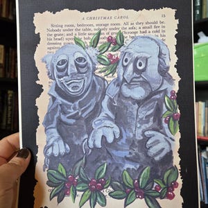 May include: A rectangular print on a page from A Christmas Carol featuring two blue-toned, puppet-like figures with large eyes. The figures are surrounded by painted green holly leaves and red berries, set against a black border on a cream background.