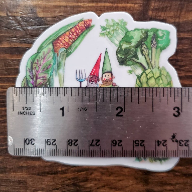 May include: A sticker featuring two gnomes with red and green hats, holding a fork and a rake, surrounded by watercolor illustrations of vegetables, including corn, broccoli, artichokes, and leafy greens. The sticker is approximately 2 inches wide.