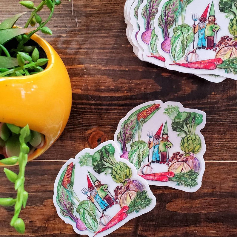 May include: Two cartoon gnomes with red hats and blue overalls are holding gardening tools in a garden scene with vegetables like artichokes, onions, carrots, and corn. The gnomes are standing on a white background with green leaves and vegetables.