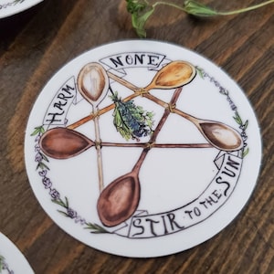 May include: A white circular sticker with a pentagram design featuring five wooden spoons arranged in a star shape. The spoons are tied together with twine. The text "NONE" is written on a banner at the top of the pentagram. The text "HARM" is written on a banner to the left of the pentagram. The text "STIR TO THE SUN" is written on a banner at the bottom of the pentagram. The sticker is surrounded by a border of small purple flowers.