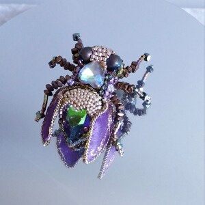 Può includere: Una spilla a forma di insetto realizzata con perline, con ali viola, gemme verdi e bianche iridescenti e dettagli marroni e dorati.