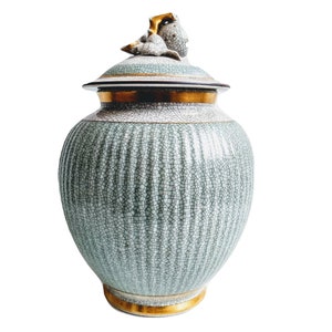 Può includere: Un vaso in ceramica verde e oro con un coperchio con un motivo floreale. Il vaso ha una superficie testurizzata e un bordo dorato.