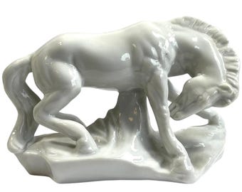 May include: A white ceramic horse figurine in a graceful pose, with its head lowered and front legs bent. The horse is mounted on a stylized base. The sculpture has a glossy finish and is a decorative item.
