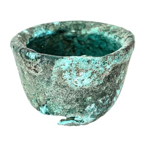 Può includere: Una piccola ciotola in ceramica di colore turchese con un bordo scheggiato. La ciotola ha una superficie ruvida e testurizzata.