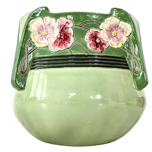 Puede incluir: Un macetero de cerámica verde con un diseño floral en relieve. Las flores son en tonos de rosa, blanco y amarillo. El macetero tiene una forma redondeada y una abertura ancha.