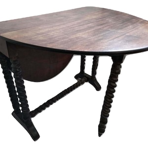 Pode incluir: Uma mesa de madeira escura com tampo dobrável e formato oval arredondado e pernas torcidas. A mesa está fechada e o tampo está para baixo.