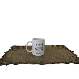 Puede incluir: Una taza de cer&aacute;mica blanca con un logotipo negro que dice "Eve Antique Pushcha" y una direcci&oacute;n de sitio web en una bandeja de metal dorado con un dise&ntilde;o ornamentado.