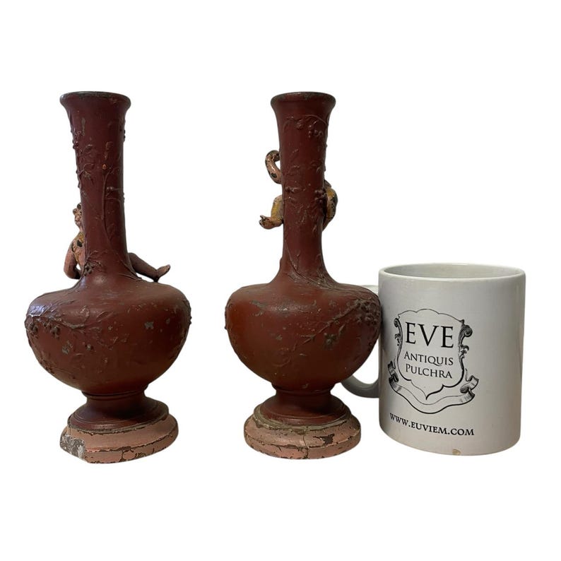 Pu&ograve; includere: Due vasi antichi color amaranto con figure di cherubini e rilievi floreali. I vasi hanno una base bulbosa e un collo lungo e sottile. Una tazza bianca con la scritta "EVE ANTIQUIS PULCHRA" &egrave; accanto ai vasi.