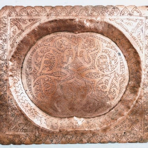 Puede incluir: Una bandeja cuadrada de cobre con un diseño intrincado en relieve. La bandeja tiene un centro elevado y redondeado y un borde decorativo.