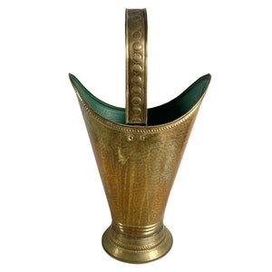 May include: A hammered brass log holder with a decorative handle. The holder has a green lining and a wide, flared base.