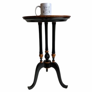 Peut inclure: Une table d'appoint en bois noir et brun avec trois pieds et un plateau rond. La table a un design simple avec un petit plateau rond et trois pieds qui s'évasent vers le bas. La table est peinte en noir avec des accents bruns sur les pieds et le plateau.