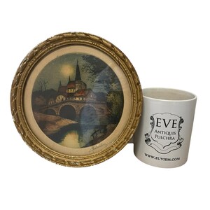Puede incluir: Una impresi&oacute;n enmarcada de una escena de un pueblo europeo con un puente y una iglesia, en un marco dorado y ornamentado. Junto a ella, una taza de cer&aacute;mica blanca con el texto "EVE ANTIQUIS PULCHRA" y una direcci&oacute;n web.