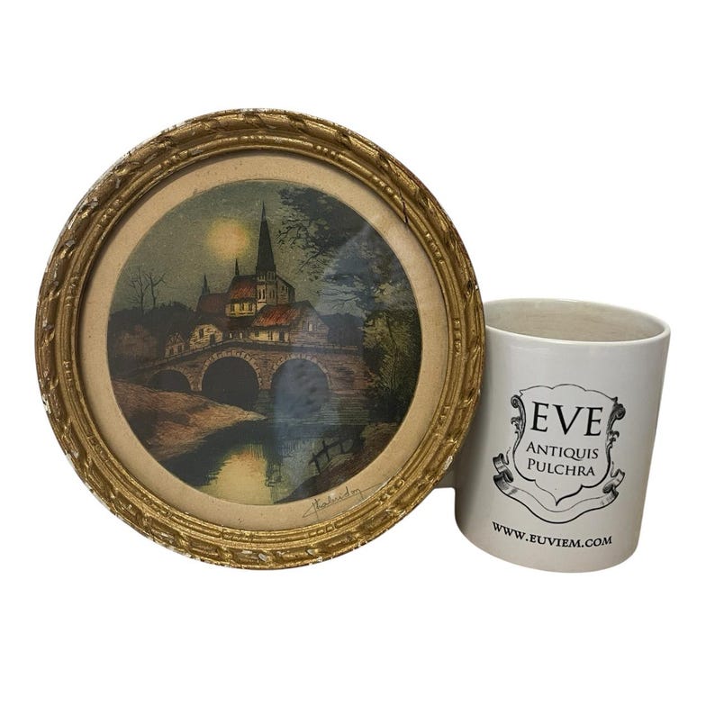 Puede incluir: Una impresi&oacute;n enmarcada de una escena de un pueblo europeo con un puente y una iglesia, en un marco dorado y ornamentado. Junto a ella, una taza de cer&aacute;mica blanca con el texto "EVE ANTIQUIS PULCHRA" y una direcci&oacute;n web.