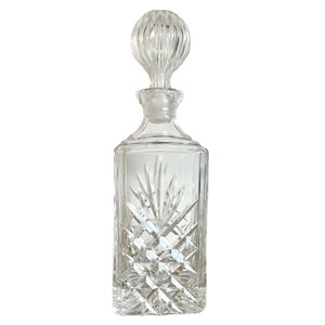 May include: A clear glass decanter with a square body and a faceted design. The decanter has a round stopper with a faceted design.