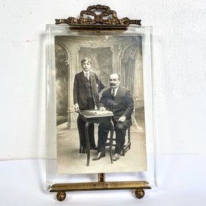May include: A vintage brass picture frame with a glass front and a stand. The frame holds a black and white photograph of two men in suits sitting at a table.