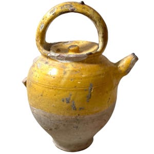May include: An antique yellow and tan ceramic teapot with a handle and spout. The teapot has a rounded body and a lid. The handle is arched, and the spout is small. The ceramic has a distressed appearance.