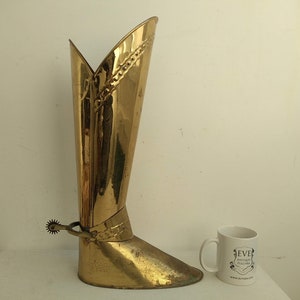 May include: A gold-colored metal boot-shaped vase with a spur detail. The vase is tall and narrow, with a pointed toe. It is a decorative item that could be used to hold flowers or other decorative items.