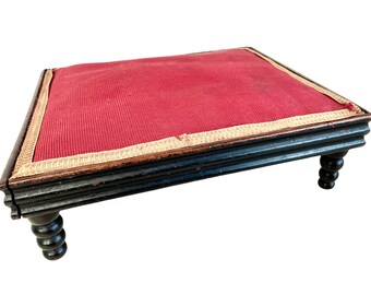 May include: A small, black wooden footstool with turned legs and a red fabric top. The fabric has a gold trim around the edges.