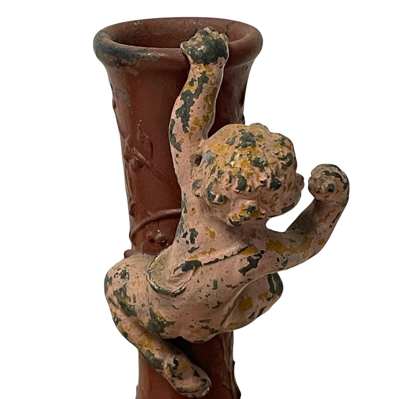 Pu&ograve; includere: Un vaso vintage marrone-rossastro con un cherubino che si arrampica sul lato. Il cherubino &egrave; dipinto in rosa sbiadito, giallo e verde, con usura visibile. Il vaso ha una superficie strutturata e un bordo scuro, suggerendo un'estetica antica o invecchiata.