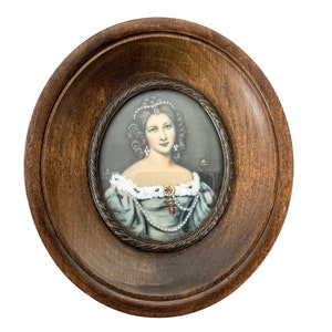Puede incluir: Un retrato enmarcado de una mujer con un vestido azul y blanco, un collar de perlas y un colgante de joyas. El retrato está enmarcado en un marco ovalado de madera oscura.