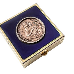 May include: A small, square, gold-colored box with a blue enamel top. The box features a round, silver-colored coin with a religious image and the inscription "NOTRE DAME DU SACRE COEUR".