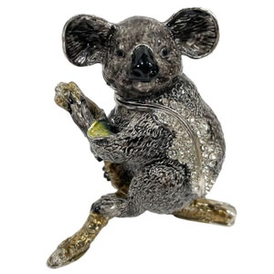 Könnte beinhalten: Eine graue Koala-Figur aus Metall mit schwarzer Nase und Augen, die auf einem Ast mit grünen Blättern sitzt. Der Koala ist mit Strasssteinen verziert.