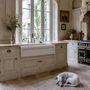 May include: A traditional kitchen featuring a white farmhouse sink, brass tap, and cream cabinets. A white dog lies on the stone floor. A large window with climbing plants provides natural light. A black stove with gold detailing is present.