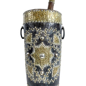 May include: A tall, cylindrical, black leather container with gold and silver accents. The container is decorated with a geometric pattern and has two metal rings on the sides. A wooden pestle sits on top of the container.