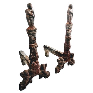 Puede incluir: Un par de soportes de chimenea de hierro fundido antiguo con un acabado marrón rústico. Cada soporte presenta una pequeña figura detallada de pie sobre una base decorativa.