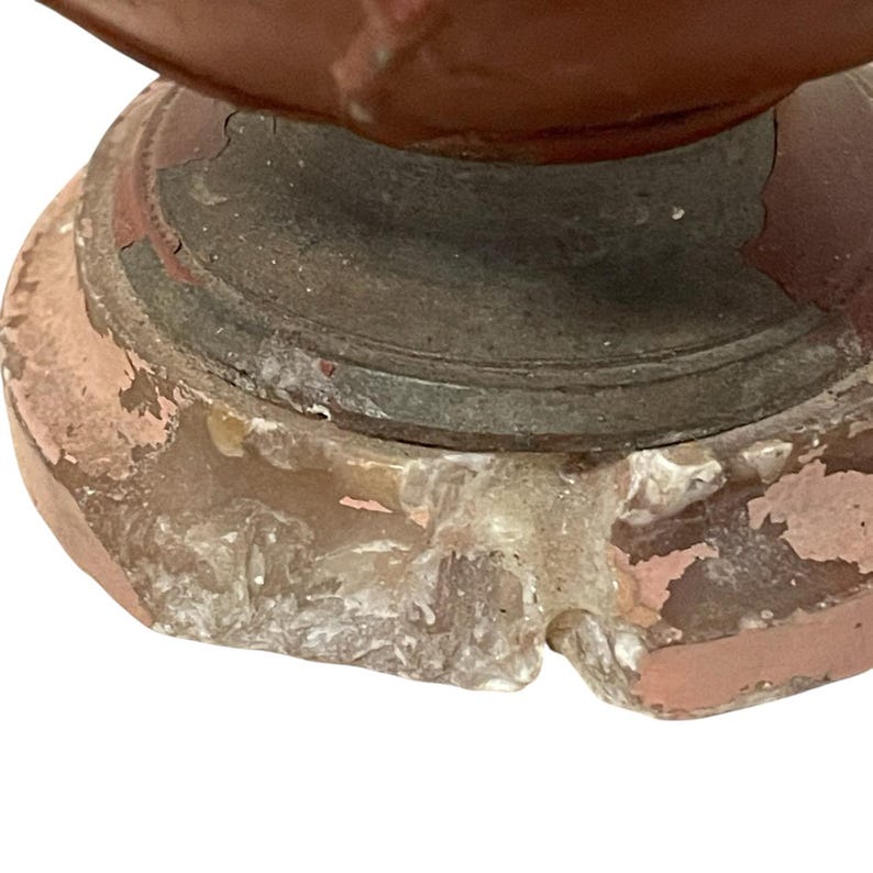 Pu&ograve; includere: Primo piano di un oggetto antico e usurato. Presenta una parte superiore marrone-rossastra e una base grigia circolare. La base ha un colore beige rosato chiaro con vernice scheggiata. L'oggetto appare invecchiato e usurato.