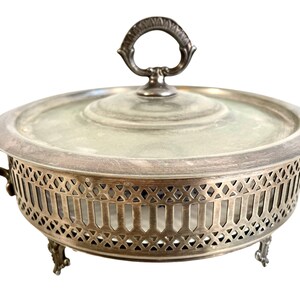 Puede incluir: Plato de servir plateado con un inserto de vidrio y una tapa. El plato tiene un diseño decorativo calado alrededor del borde y tres patas.