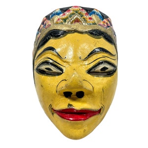 May include: A yellow wooden mask with a painted face, black eyebrows, and red lips. The mask has a colourful crown with gold accents.