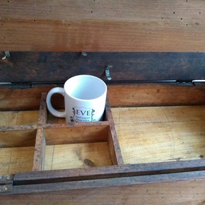 May include: A wooden box with a hinged lid and a divided interior. The box is open and a white ceramic mug with the text "Eve Antiquit&eacute;s Chic & Rare" is sitting inside.