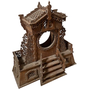 May include: A detailed, brown wooden architectural model with intricate carvings. Features include a circular window, stairs, and a decorative roof. The model has a rustic, aged appearance, suggesting it is vintage or antique.