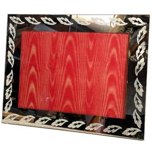 May include: A silver-toned picture frame with a black border and a red velvet backing. The frame has a decorative design of silver leaves around the border.