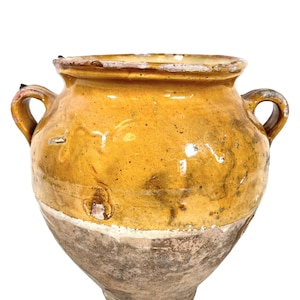 May include: A large, yellow glazed earthenware pot with two handles. The pot has a rustic, aged appearance with a brown base.