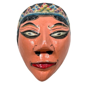 Peut inclure: Un masque en bois peint à la main avec un visage rose, des yeux noirs et une bouche rouge. Le masque a une couronne bleue, jaune et verte sur le dessus.