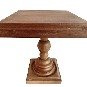 May include: A wooden pedestal cake stand with a square top and a turned pedestal base. The stand is made of natural wood with a light brown finish.