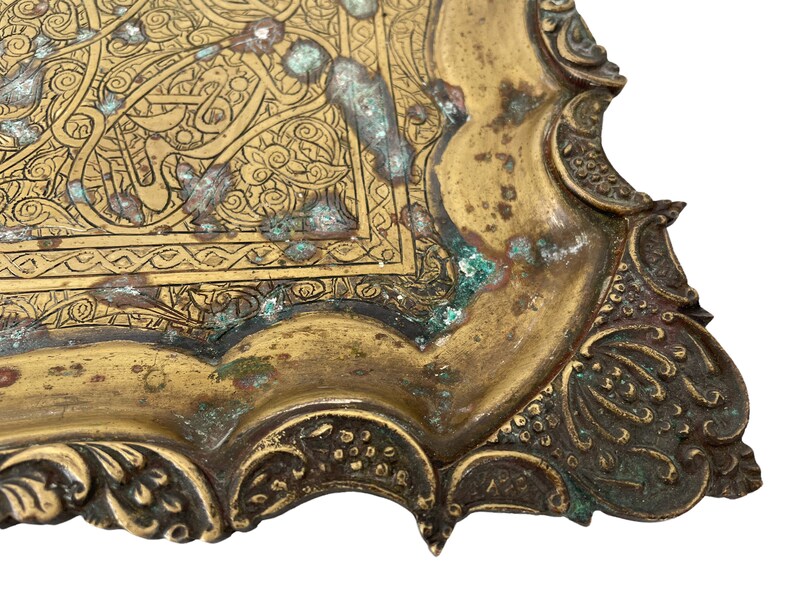 Puede incluir: Una bandeja de metal de color dorado con un dise&ntilde;o grabado intrincado. La bandeja tiene un borde festoneado y est&aacute; decorada con motivos florales y geom&eacute;tricos. La bandeja muestra signos de edad y desgaste, con algunas &aacute;reas de deslustre y p&aacute;tina.