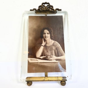 May include: A vintage glass picture frame with a gold metal stand and ornate design. The frame holds a black and white photograph of a woman with dark hair, wearing a crocheted top and holding a book.