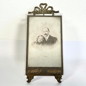 May include: A vintage brass picture frame with a glass front and a decorative bow at the top. The frame has a black and white photo of a man and a woman inside. The frame has the text "R. Wingend" and "Borghorst a.d.M" on the bottom.