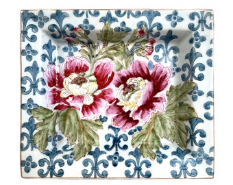May include: A square ceramic dish with a white background and a blue and gold floral pattern. The dish features two large pink flowers with yellow centers and green leaves.