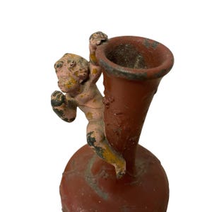 Pu&ograve; includere: Un piccolo vaso in stile antico nei toni del rosso e del rosa, con una figura di cherubino aggrappata al lato. Il vaso ha una parte superiore svasata e una base arrotondata. Il cherubino &egrave; dipinto di rosa con accenti gialli e neri.